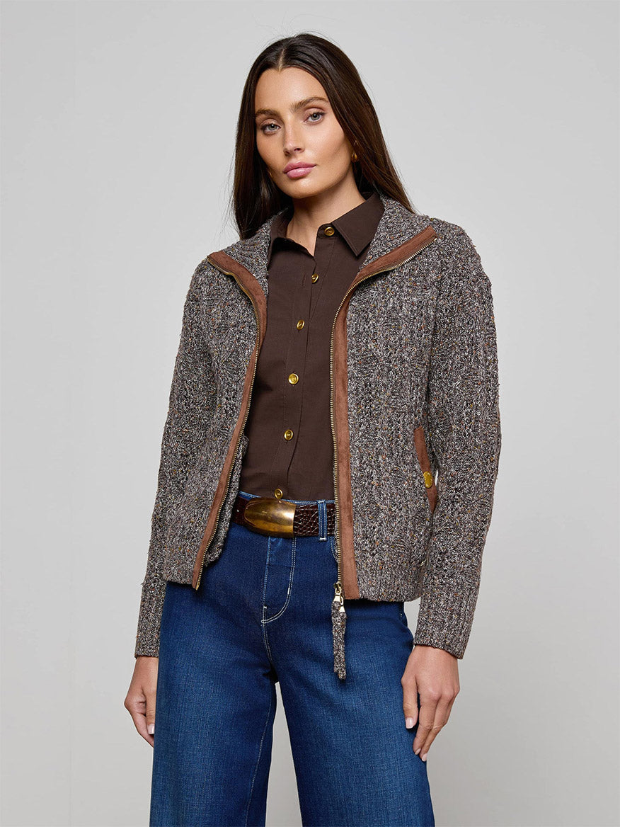 Woman standing against a plain background, wearing the L'Agence Shiloh Cable Knit Cardigan in Carafe Brown Multi, paired with blue jeans and a brown belt.