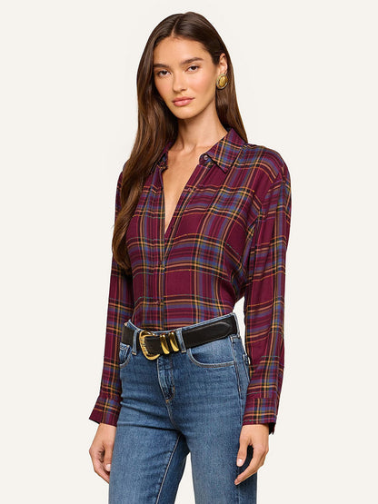 A woman wearing the L’Agence Suzette Shirt in Dark Plum Multi Plaid—a long-sleeve, deep V-neck shirt with a high-low hemline—tucked into blue jeans with a black belt, stands against a plain background.