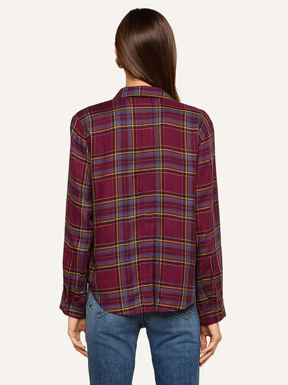 A person with long brown hair stands facing away, wearing the L’Agence Suzette Shirt in Dark Plum Multi Plaid with a high-low hemline and blue jeans against a plain, light background.