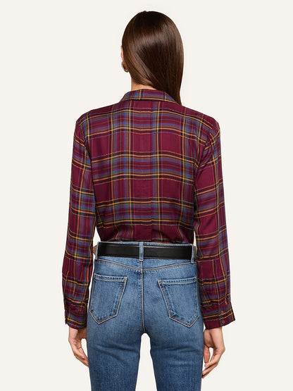 A person with straight brown hair is shown from the back, wearing the L’Agence Suzette Shirt in Dark Plum Multi Plaid tucked into blue jeans with a black belt. The button-down shirt features classic plaid patterning that adds a timeless touch to the casual outfit.