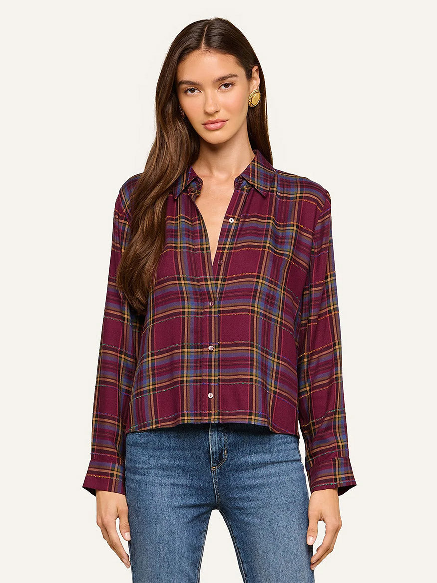 A woman wearing the L’Agence Suzette Shirt in Dark Plum Multi Plaid—featuring long sleeves, a button-down front, and a high-low hemline—is paired with blue jeans and stands against a plain light background.