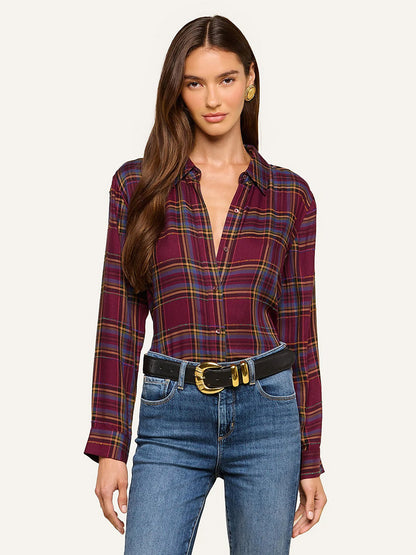 A woman wearing the L’Agence Suzette Shirt in Dark Plum Multi Plaid, featuring long sleeves, button-down front, and a chic high-low hemline, styled with blue jeans and a black belt with a gold buckle, stands against a neutral background.