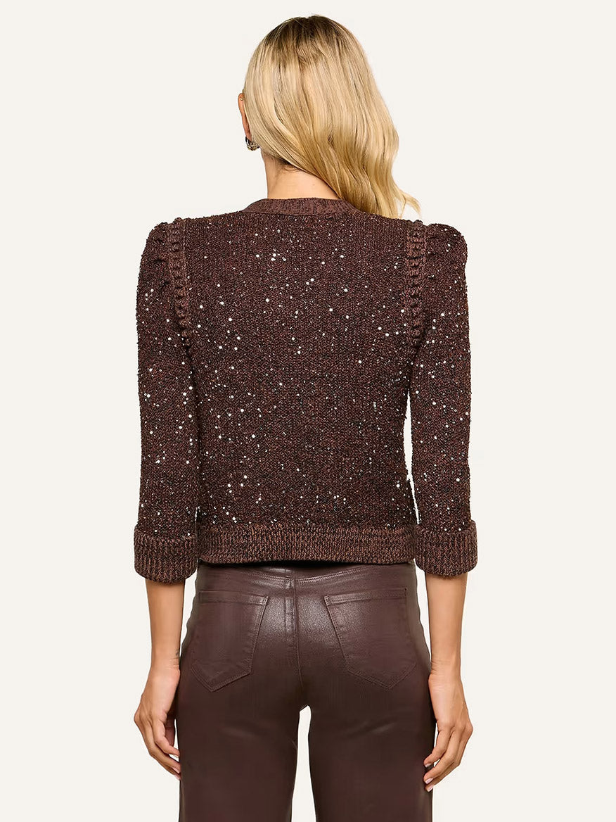 A woman with long blonde hair is seen from the back wearing the L’Agence Thistle Sequined Cardigan in Black/Java/Sequins, featuring micro sequin embellishments and puffed shoulders, paired with brown leather pants—a sophisticated look for Fall 2026.