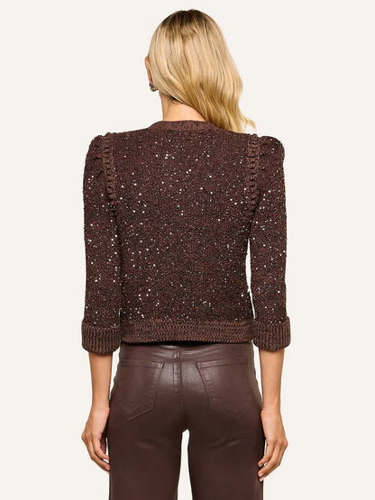 A woman with long blonde hair is seen from the back wearing the L’Agence Thistle Sequined Cardigan in Black/Java/Sequins, featuring micro sequin embellishments and puffed shoulders, paired with brown leather pants—a sophisticated look for Fall 2026.