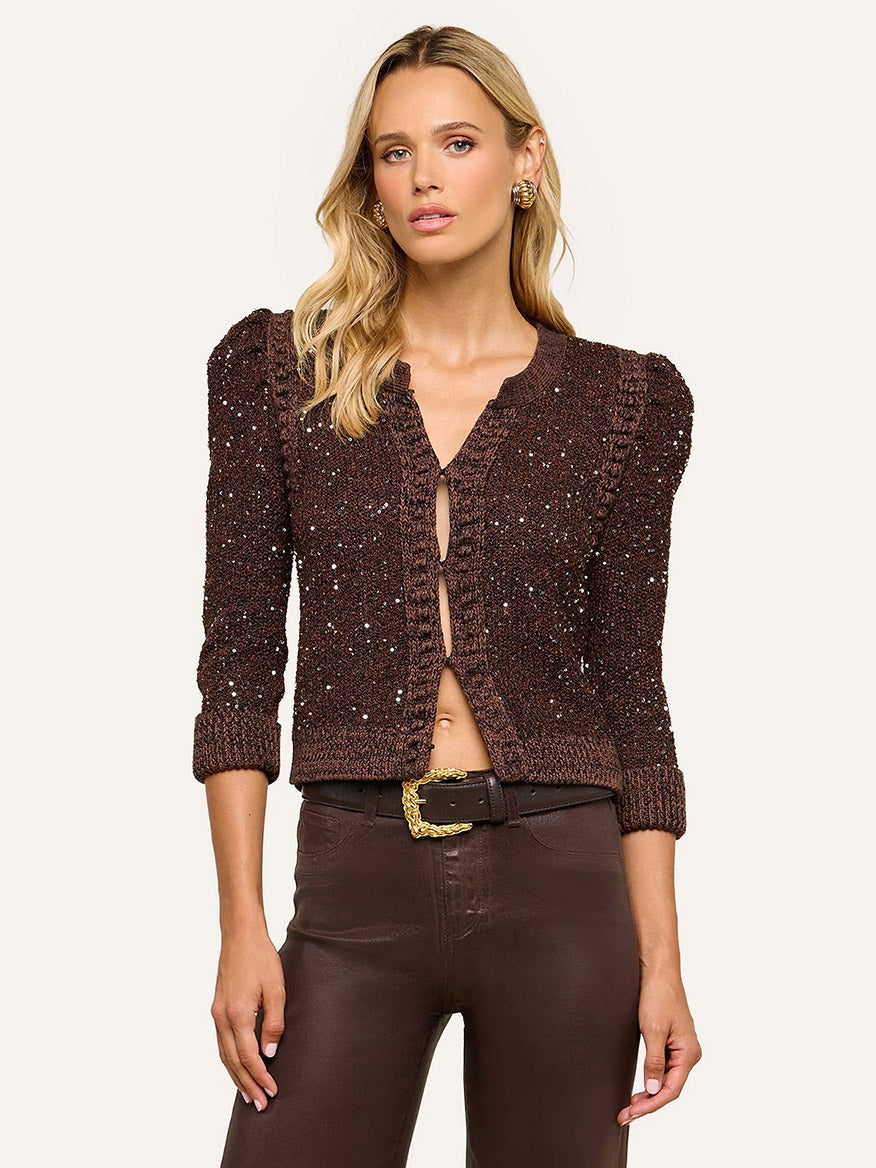 A woman wearing the L’Agence Thistle Sequined Cardigan in Black/Java/Sequins, featuring textured fabric, puffed shoulders, and subtle micro sequins, paired with brown high-waisted pants and standing against a plain background—an elegant look perfect for Fall 2026.