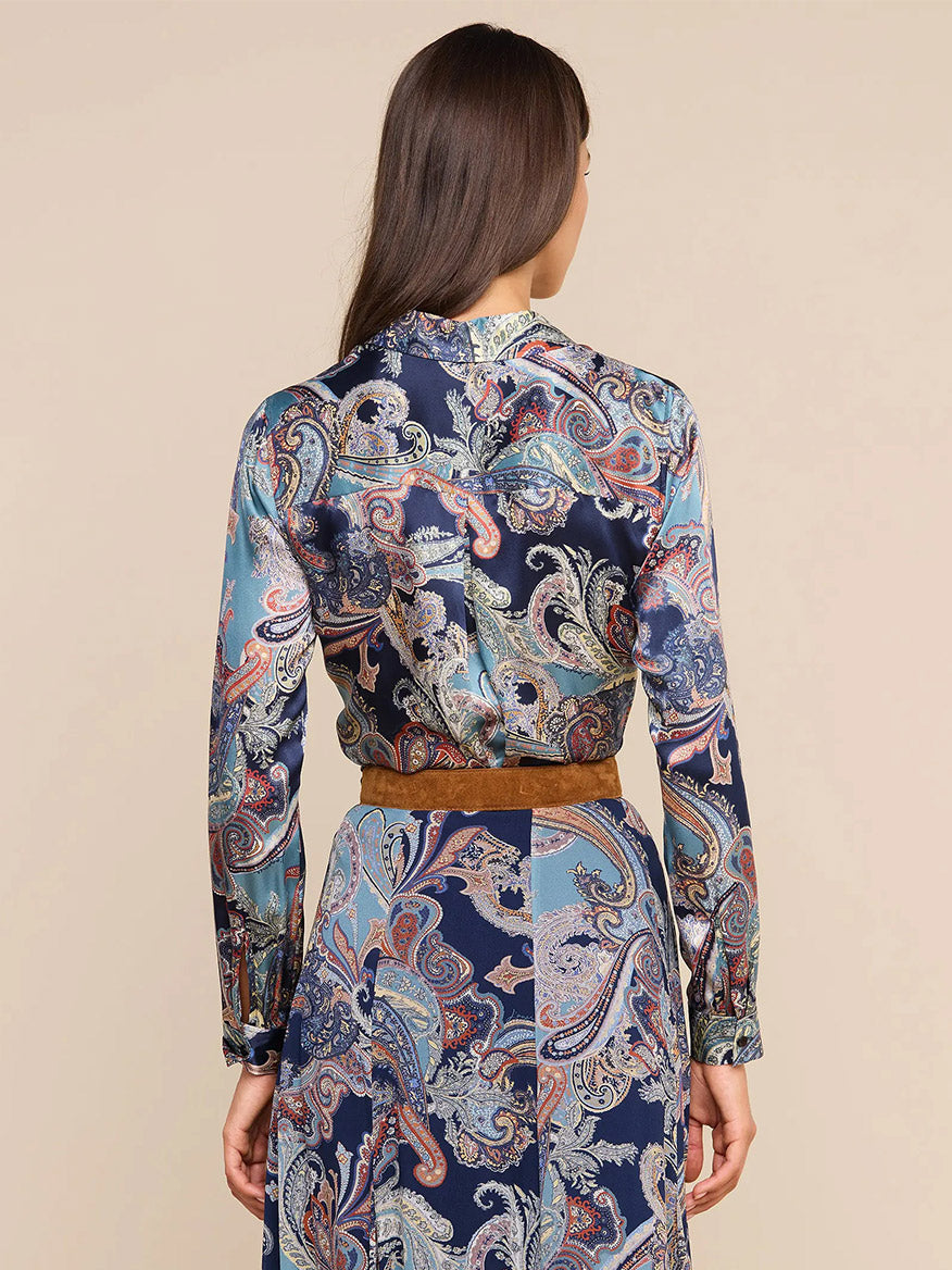 A woman stands facing away, wearing the L’Agence Tyler Blouse in Midnight Multi Besarabian Paisley—a long-sleeve silk button-down adorned with an elegant paisley print in shades of blue, brown, and cream—paired with a matching skirt and finished with a brown belt at the waist.