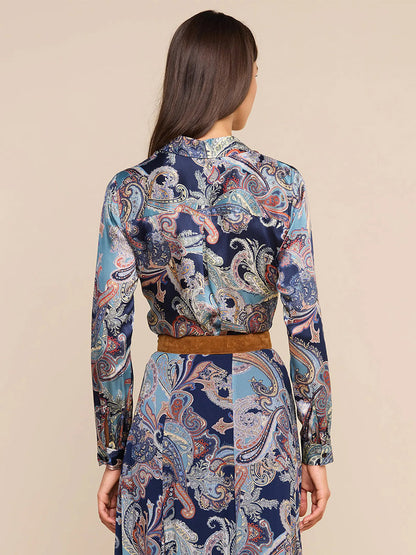 A woman stands facing away, wearing the L’Agence Tyler Blouse in Midnight Multi Besarabian Paisley—a long-sleeve silk button-down adorned with an elegant paisley print in shades of blue, brown, and cream—paired with a matching skirt and finished with a brown belt at the waist.