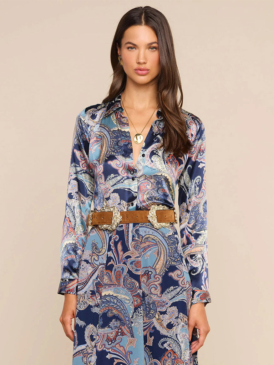 Woman wearing the L’Agence Tyler Blouse in Midnight Multi Besarabian Paisley, featuring long sleeves and a paisley print in blue and purple hues, styled with a gold necklace and a wide brown belt with large gold buckles.