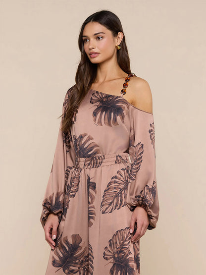 Woman wearing the L'Agence Asami Chain One-Shoulder Blouse in Hazelnut Multi Large Palm, featuring a palm print, standing against a plain beige background.