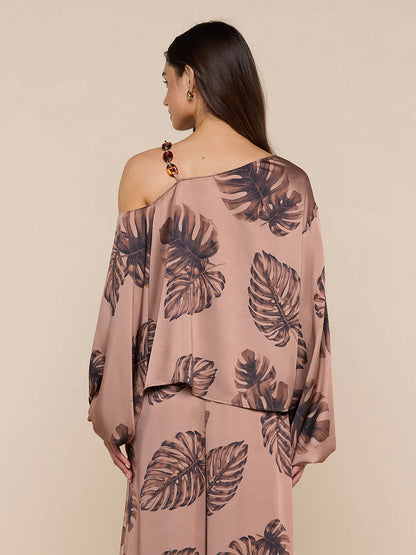 A woman with long brown hair is seen from the back wearing the L'Agence Asami Chain One-Shoulder Blouse in Hazelnut Multi Large Palm, featuring a satin finish and large palm print.