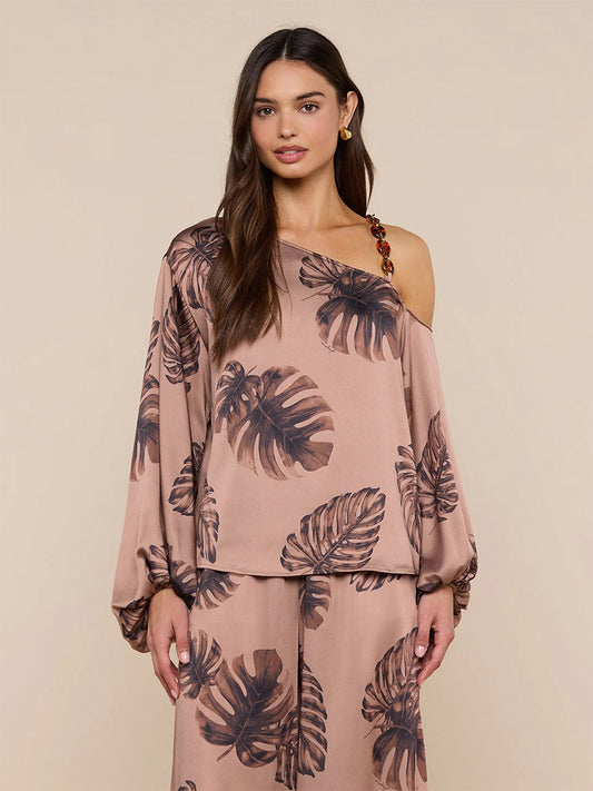 Woman wearing the L'Agence Asami Chain One-Shoulder Blouse in Hazelnut Multi Large Palm, paired with matching pants featuring a brown palm print, stands against a plain beige background.