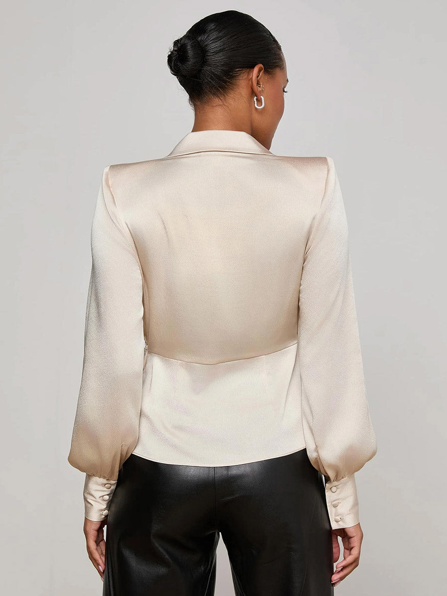 A woman stands with her back to the camera, wearing the L'Agence Bernice Satin Wrap Blouse in Champagne, styled with long sleeves and paired with black leather pants for a look of romantic elegance.