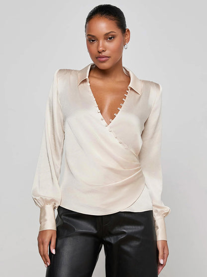 A woman stands against a plain background wearing the L'Agence Bernice Satin Wrap Blouse in Champagne with long sleeves and black leather pants, exuding romantic elegance.