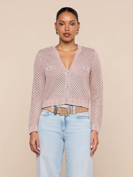 A woman stands against a beige background wearing the L'Agence Blanca Sequin Cardigan in Adobe Rose—a light pink sequined knit with button details—paired with light blue jeans and a beige belt.