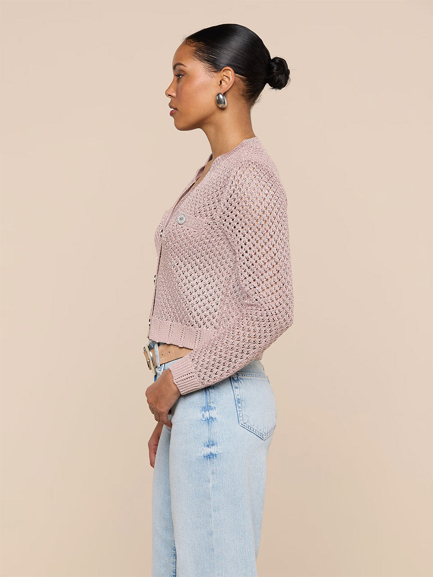 A woman stands in profile wearing the L'Agence Blanca Sequin Cardigan in Adobe Rose with light blue jeans against a plain beige background, showcasing this chic lightweight layer.