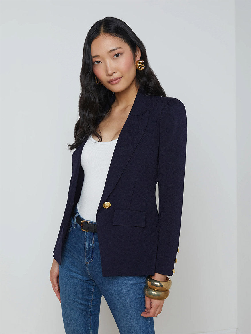 A woman with long dark hair wears the L'Agence Chamberlain Knit Blazer in Midnight/Gold with embossed dome buttons, a white top, blue jeans, and gold hoop earrings while standing against a plain background.