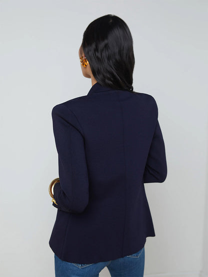 A person with long black hair stands with their back to the camera, wearing a L'Agence Chamberlain Knit Blazer in Midnight/Gold adorned with embossed dome buttons and blue jeans.