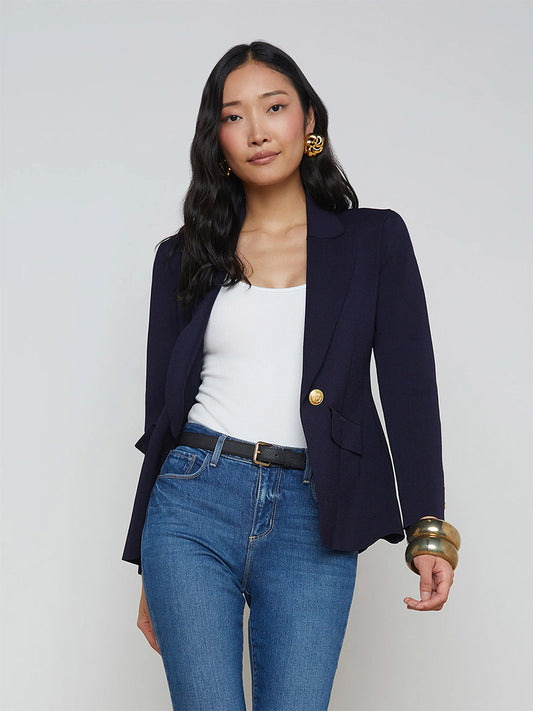 A woman with long black hair is wearing a white top, the L'Agence Chamberlain Knit Blazer in Midnight/Gold with embossed dome buttons, blue jeans, and gold accessories. She stands against a plain background.