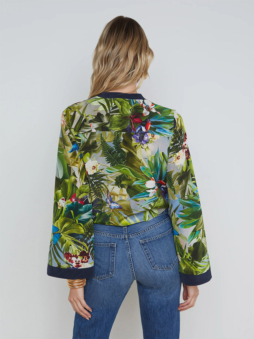 A person with long, wavy hair is seen from the rear, wearing a floral-patterned jacket, blue jeans, and the L'Agence Charlize Tie-Front Silk Blouse in Multi Lush Jungle with dramatic bell sleeves. They are standing against a plain background.