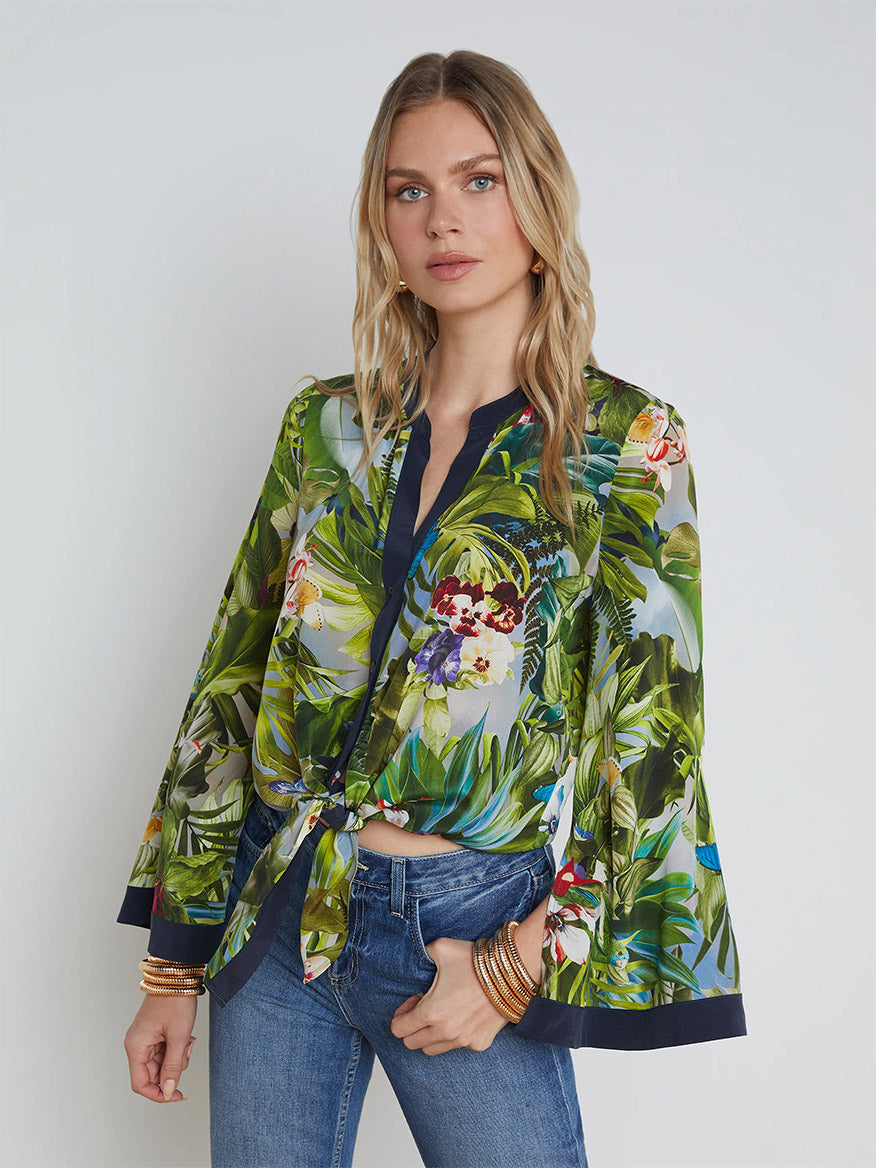 A woman with long blonde hair stands against a plain background, wearing the L'Agence Charlize Tie-Front Silk Blouse in Multi Lush Jungle—an elegant 100% silk blouse featuring dramatic bell sleeves and a tie waist—paired with blue jeans.