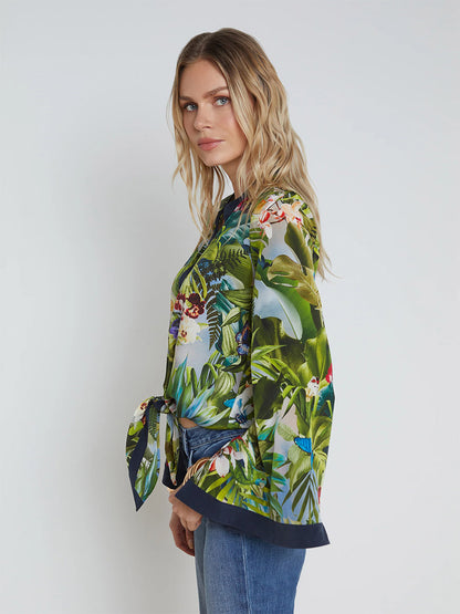 A woman with wavy blonde hair, wearing the L'Agence Charlize Tie-Front Silk Blouse in Multi Lush Jungle paired with blue jeans, stands against a plain white background, looking at the camera.