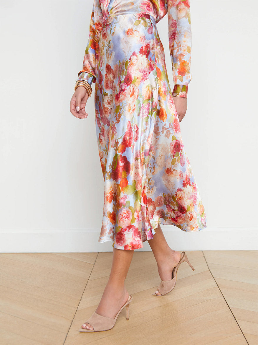A person wearing a L'Agence Clarisa Silk Skirt in Multi Soft Cloud Floral and beige high-heeled sandals, standing on a wooden floor.
