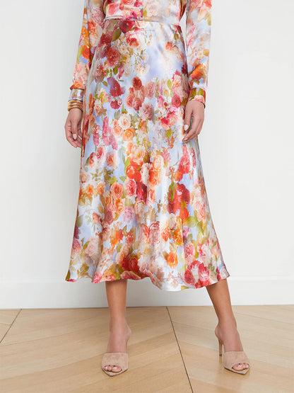 A person wearing a L'Agence Clarisa Silk Skirt in Multi Soft Cloud Floral and beige heeled sandals, standing on a wooden floor against a white background.