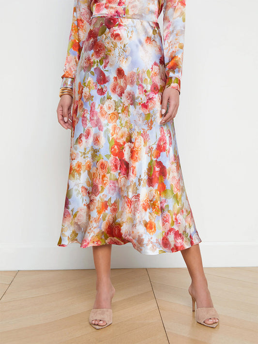 A person wearing a L'Agence Clarisa Silk Skirt in Multi Soft Cloud Floral and beige heeled sandals, standing on a wooden floor against a white background.