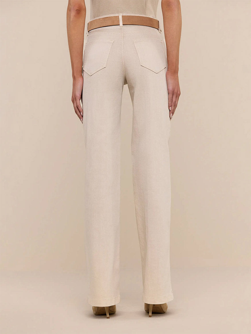 A person stands facing away from the camera, wearing L'Agence Clayton Wide-Leg Jean in Biscuit paired with a brown belt and beige high-heeled shoes against a plain background.