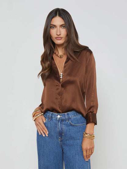 A woman with long hair models the iconic L'Agence Dani Blouse in Hot Fudge, paired with blue jeans and accented by gold jewelry, against a simple backdrop.