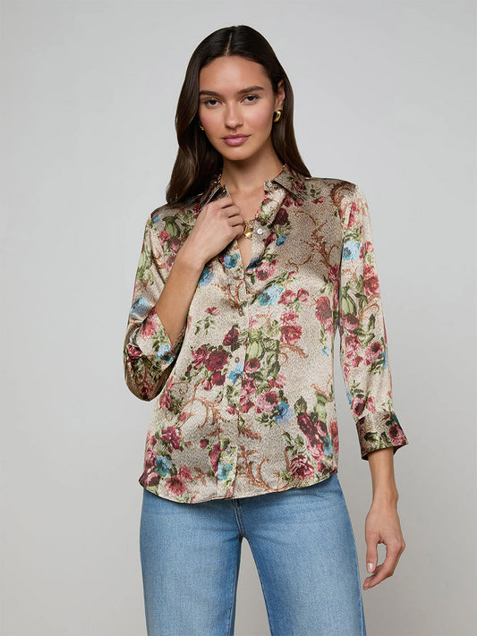 A woman wears the L'Agence Dani Blouse in Multi Floral Tapestry with long sleeves and blue jeans, standing against a plain background.