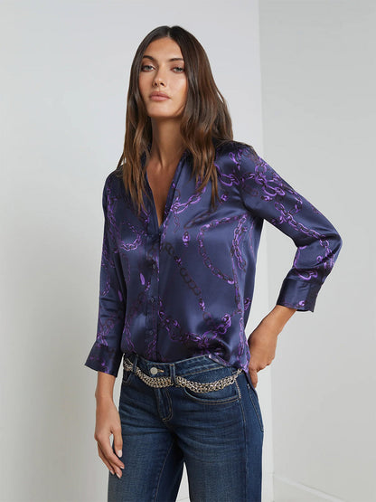 A woman wearing the L'Agence Dani Blouse in Midnight/Purple Sketch Chain paired with jeans and a chain belt is standing against a light gray background.