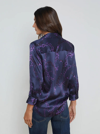 The individual with long hair is donning the L'Agence Dani Blouse in Midnight/Purple Sketch Chain, featuring a dark, three-quarter sleeve design with purple swirls. It adds an elegant touch to the ensemble and is showcased from the back against a plain background.