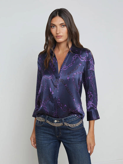 A woman stands against a plain background, wearing the L'Agence Dani Blouse in Midnight/Purple Sketch Chain with three-quarter sleeves, paired with jeans and a chain belt.