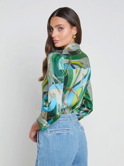 A woman in a L'Agence Dani Blouse in Sea Green Multi Belt and blue jeans looks over her shoulder against a plain background.