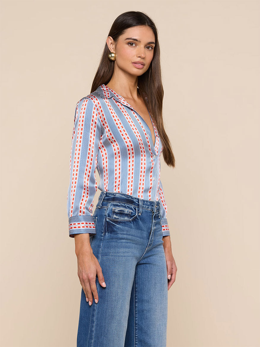 A woman stands against a beige background, wearing the L'Agence Dani Blouse in Blue/Red Textured Stripe Print—a long-sleeve silk button-down—paired with high-waisted blue jeans.