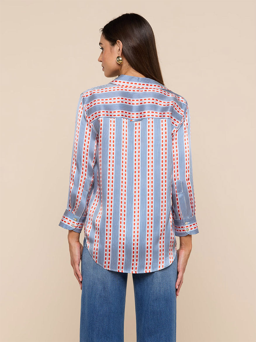 A woman is seen from the back wearing the L'Agence Dani Blouse in Blue/Red Textured Stripe Print with blue jeans, set against a plain beige background.