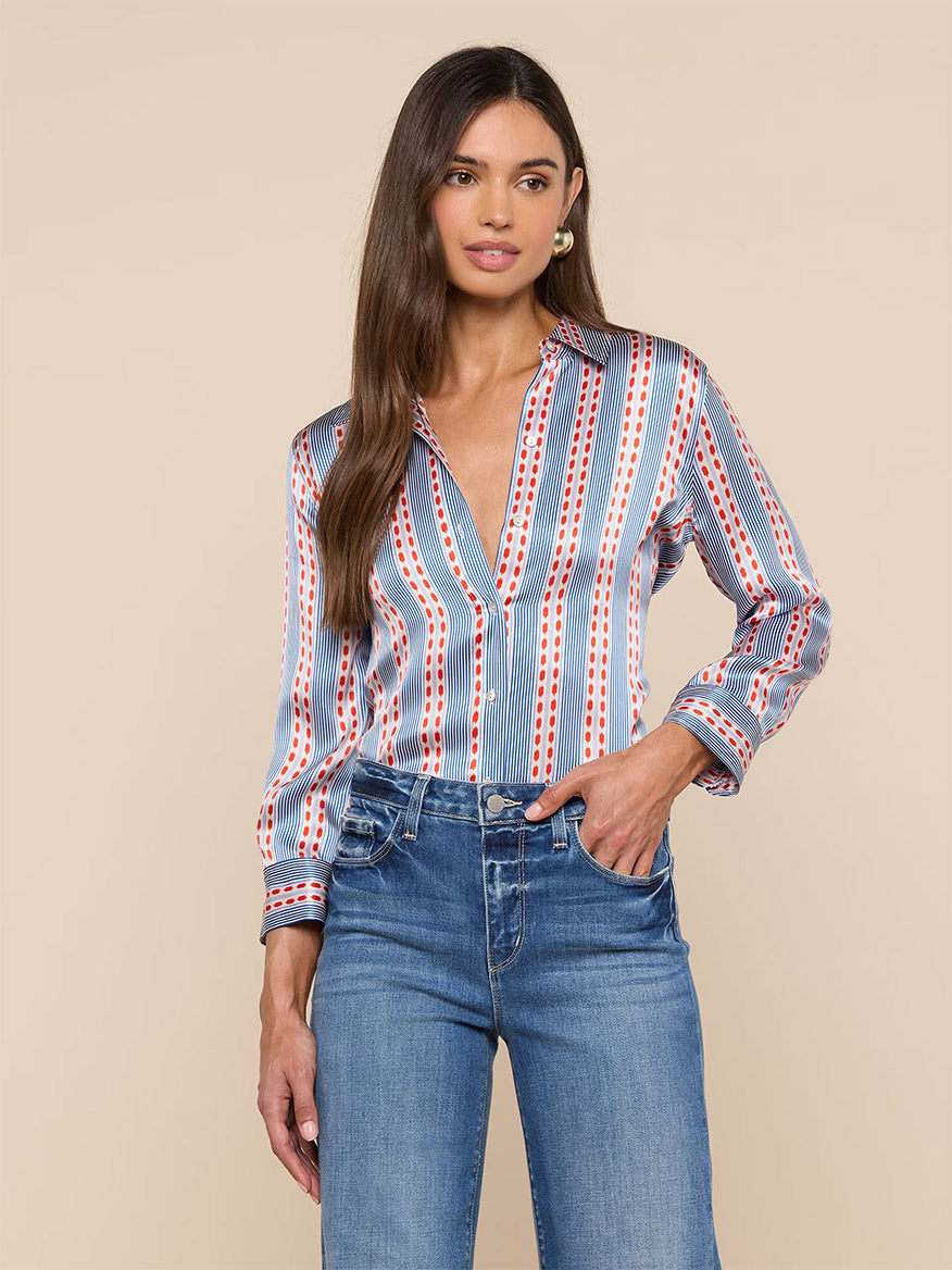 A woman wears the L'Agence Dani Blouse in Blue/Red Textured Stripe Print with high-waisted blue jeans, standing against a beige background.