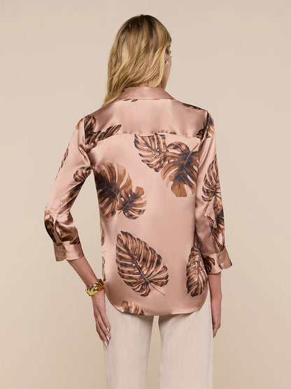 A woman is seen from the back in the L'Agence Dani Blouse in Hazelnut Multi Large Palm and cream pants, standing against a plain beige background.
