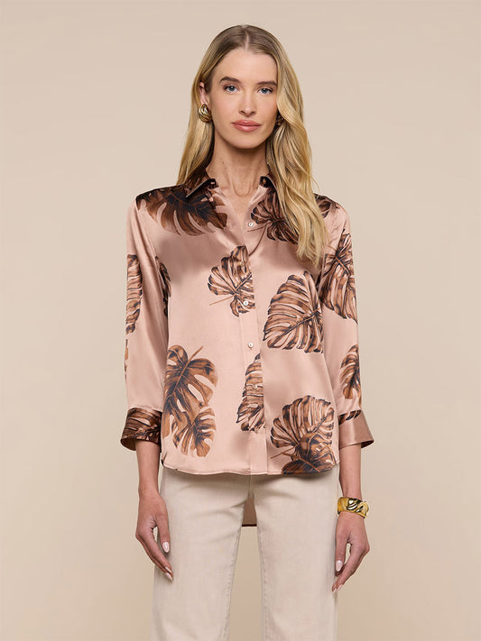 Woman stands against a beige background wearing the L'Agence Dani Blouse in Hazelnut Multi Large Palm—a shiny, light brown silk button-down with a dark palm leaf print—paired with light pants and gold accessories.