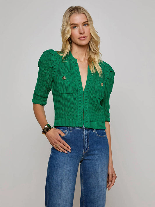 A woman wears the L'Agence Delilah Cropped Cardigan in Kelly Green/Gold Knot with short sleeves and front pockets, paired with blue jeans, standing against a plain light gray background.