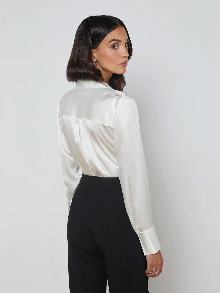 A woman in the L'Agence Divya Silk Shirt in white and black pants is looking over her shoulder, the satin-covered button closures shining subtly against a plain background.