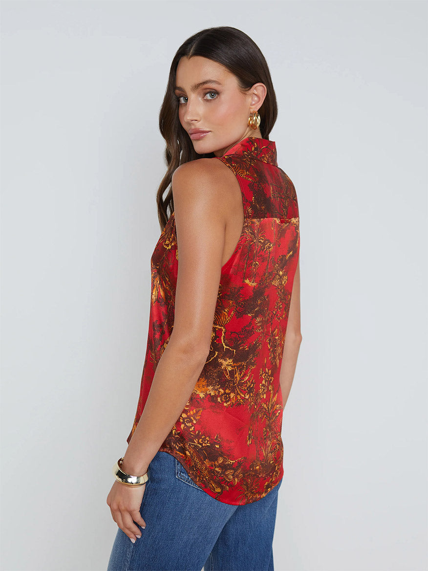 Woman with long dark hair wearing a vibrant L'Agence Emmy Sleeveless Blouse in High Risk Red Multi Jungle Toile and blue jeans, looks over her shoulder.