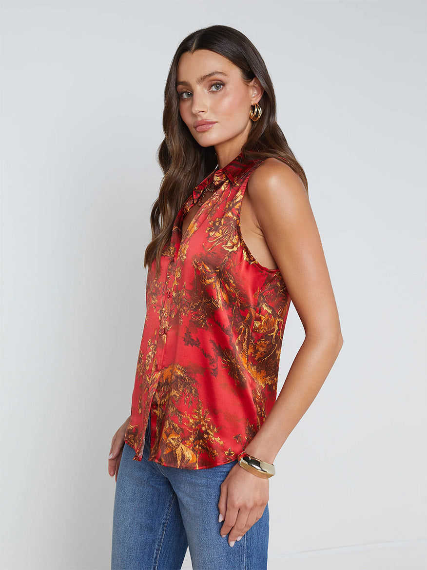 A woman with long brown hair, wearing the L'Agence Emmy Sleeveless Blouse in High Risk Red Multi Jungle Toile along with blue jeans, stands in front of a white background, looking to her left.