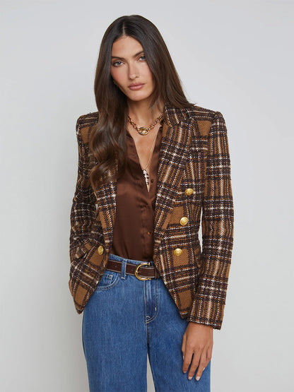 A woman in a L'Agence Kenzie Blazer featuring a blue, brown, and yellow plaid design with gold buttons, a brown blouse, and blue jeans stands against a plain background.