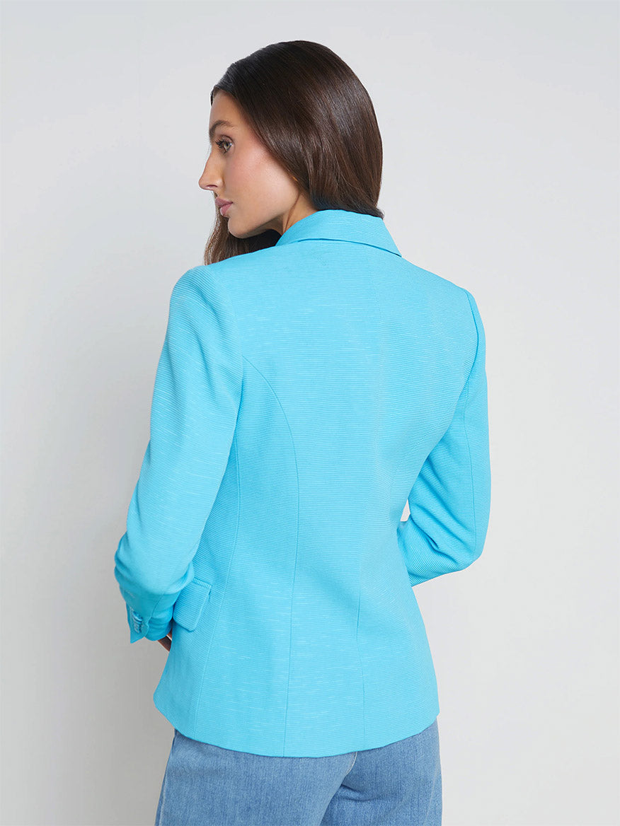 A woman viewed from the back, wearing a turquoise double-breasted L'Agence Kenzie Blazer in Blue Atoll/Multi Belt and blue jeans, against a plain white background.