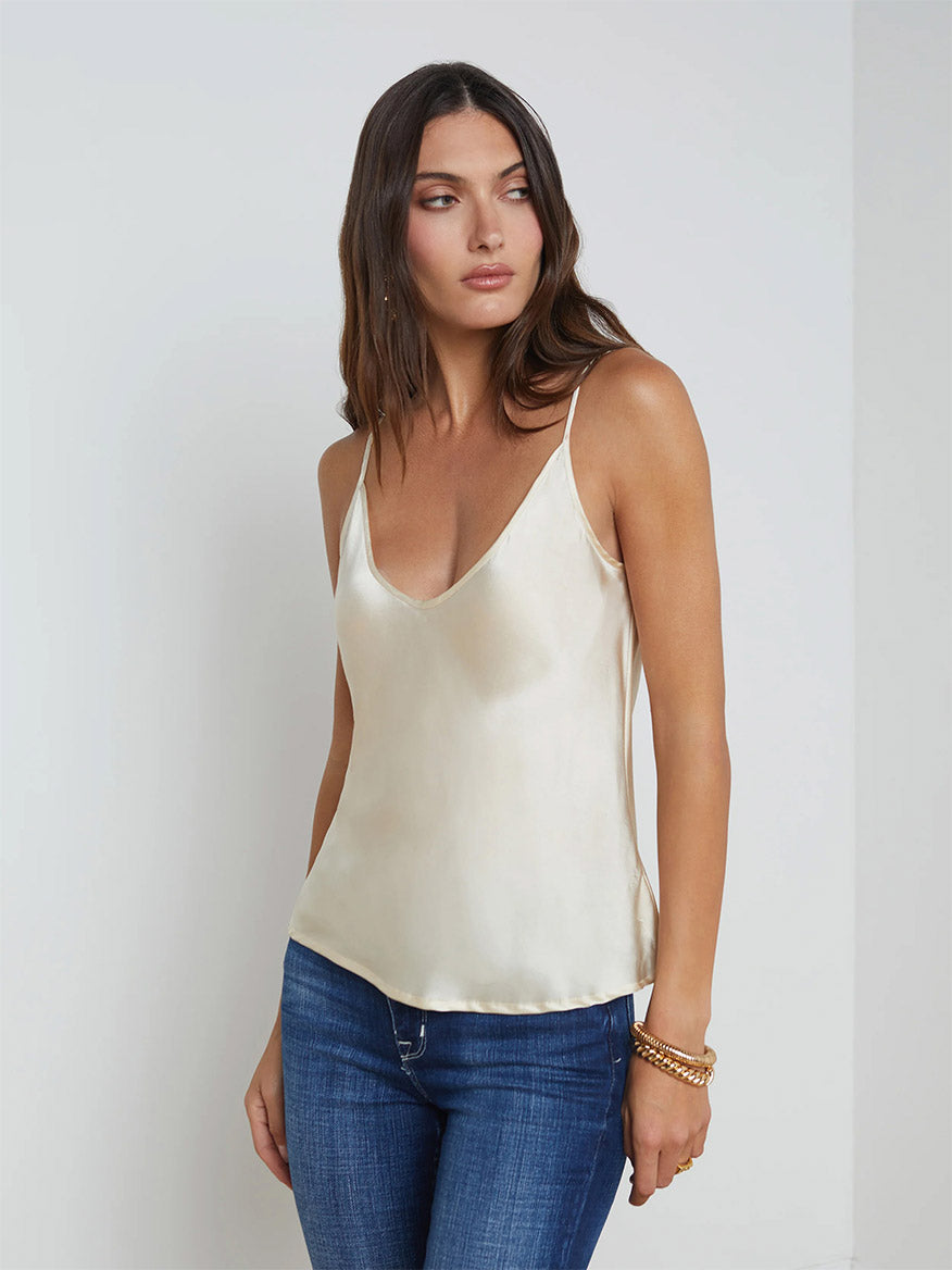 A woman in a loose L'Agence Lexi Silk Camisole in ivory with adjustable straps and blue jeans stands against a plain light-colored background, looking to her left. She is accessorized with bracelets on her right wrist, creating the perfect wardrobe foundation.