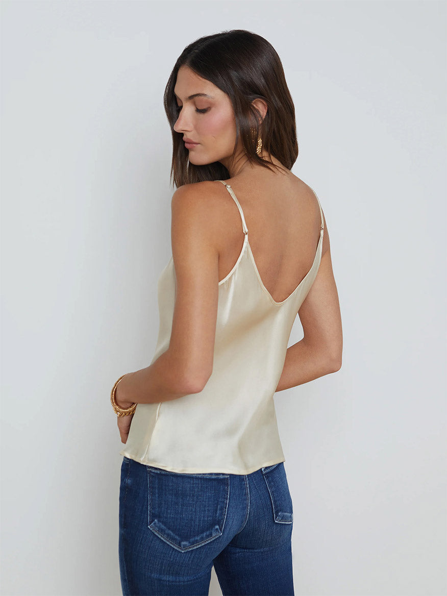 A woman with long, brown hair is wearing the L'Agence Lexi Silk Camisole in Ivory with adjustable straps and jeans, facing slightly away from the camera. She is accessorized with hoop earrings and bracelets, showcasing a timeless wardrobe foundation.