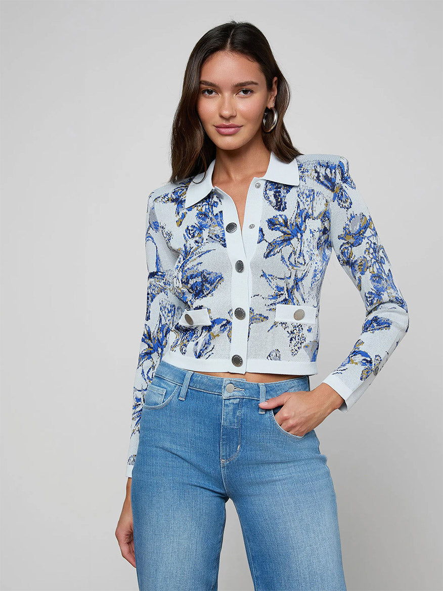 Woman wearing the L'Agence Osiris Jacquard Cardigan in Grey Multi Watercolor Butterfly Jacquard with silver snap buttons and long sleeves, paired with light blue jeans, standing against a plain background.