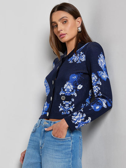 A woman stands against a gray background wearing the L'Agence Osiris Jacquard Cardigan in Black/Blue Large Porcelain, featuring long sleeves, a button-up front, a porcelain floral motif, flap pockets, and paired with light blue jeans.